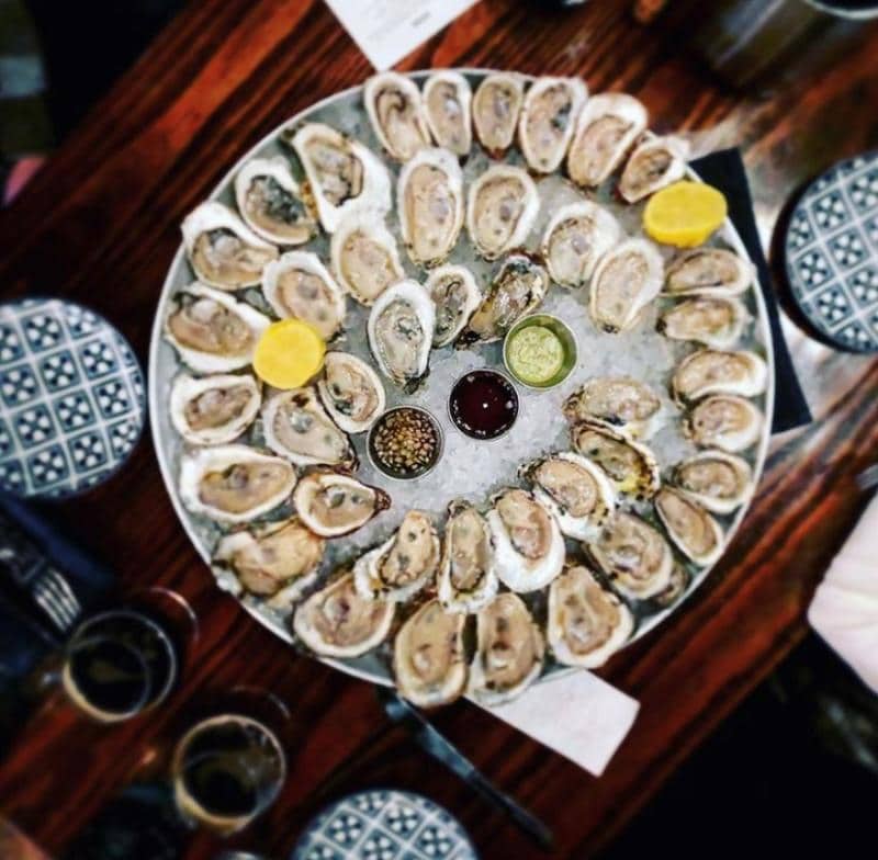A large platter of oysters on ice is surrounded by lemon wedges, dipping sauces, and seasoning, set on a wooden table with patterned plates visible at the edges.