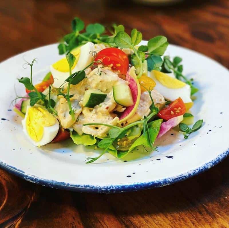 Salad with mixed greens, tomatoes, radishes, cucumber, and sliced boiled eggs on a white plate with a blue rim, placed on a wooden table.