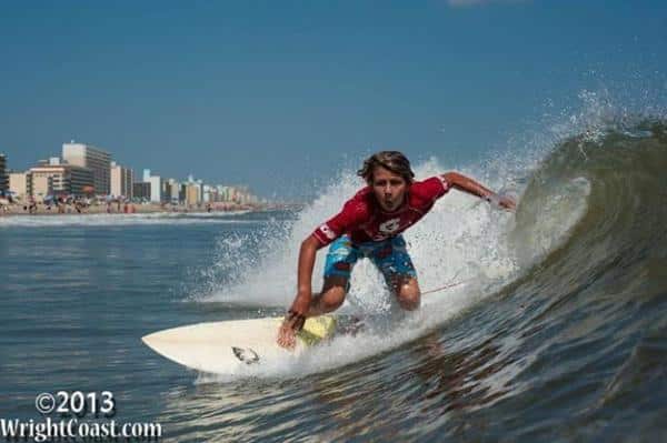 Surfer in red shirt rides a wave near a sandy beach with city buildings in the background.