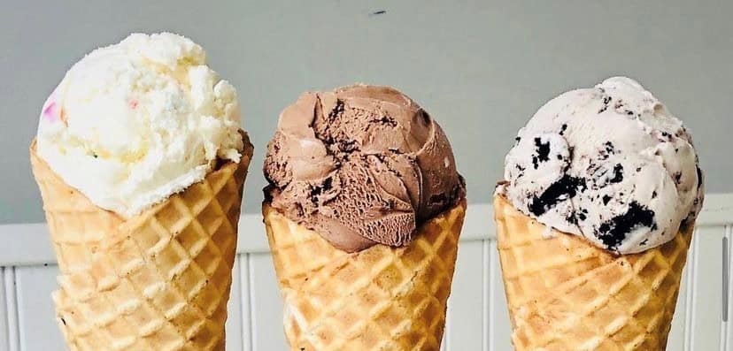Three ice cream cones on a table, featuring vanilla, chocolate, and cookies and cream flavors in waffle cones.