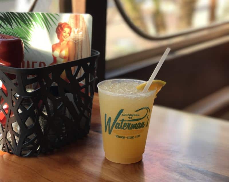 A plastic cup with an orange drink and a straw on a wooden table next to a basket containing condiments and a retro-style card.