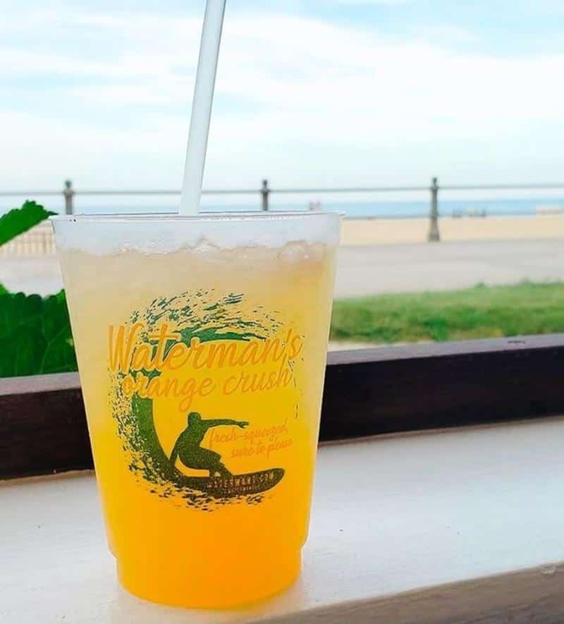 A plastic cup of orange drink with a straw on a ledge, labeled "Waterman's Orange Crush," against a beach background with a railing and ocean.