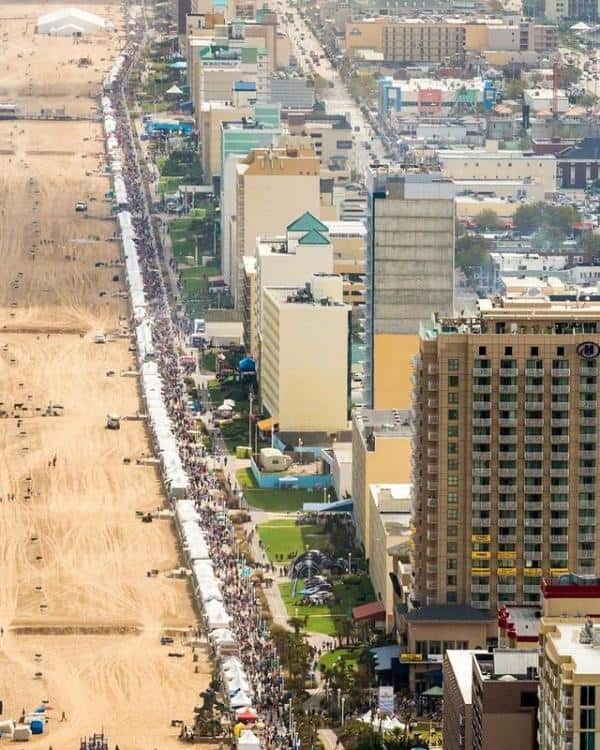 Aerial view of a long row of white tents aligned parallel to a sandy beach, with tall buildings and a busy street nearby. People walk along the tents.