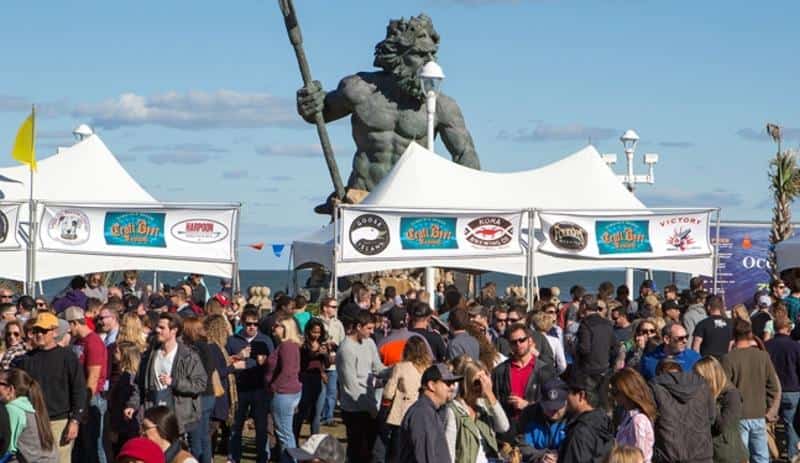 Crowd gathered at an outdoor festival with a large statue of a figure holding a staff in the background under white tents displaying various beer logos.
