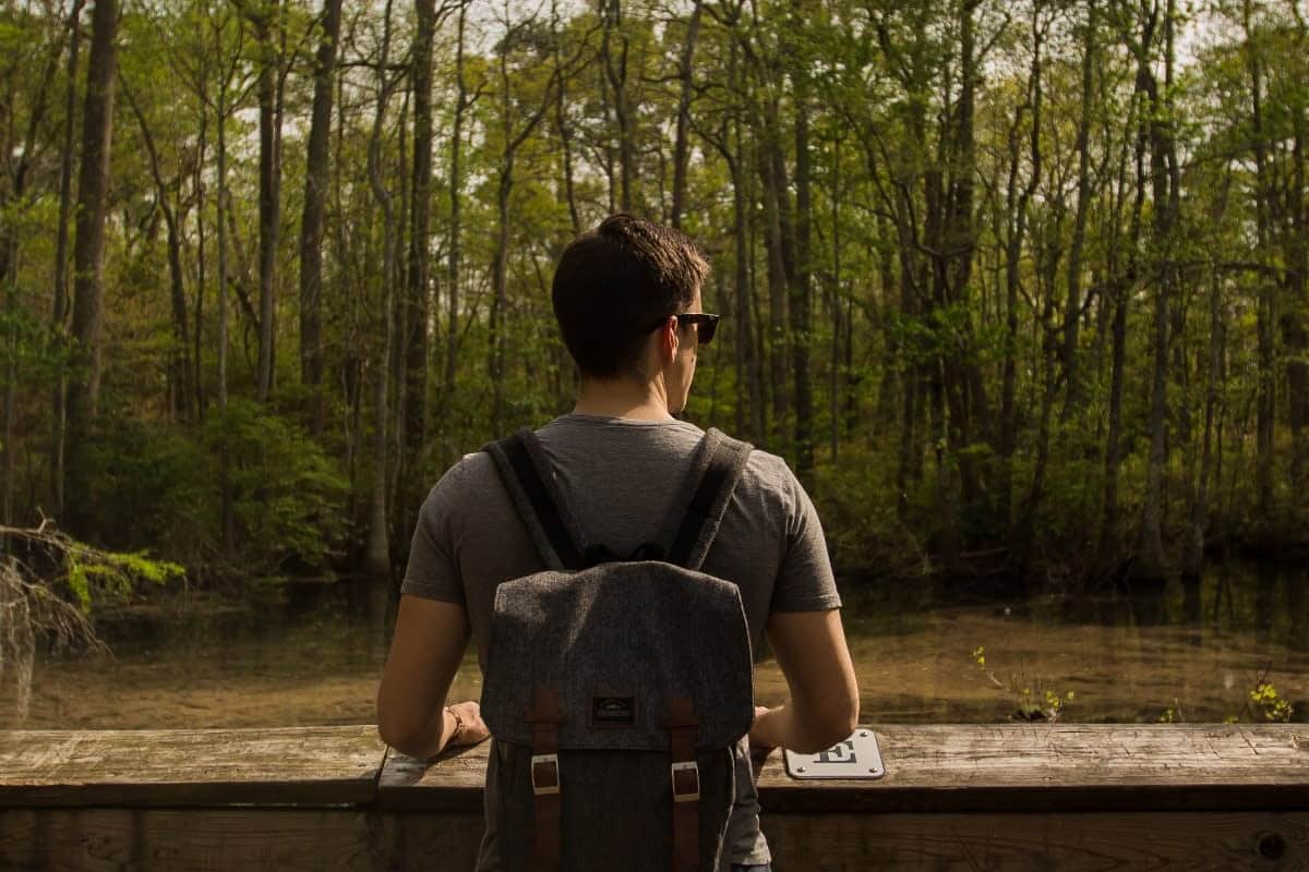 A person wearing sunglasses and a backpack stands facing a forested area, overlooking a body of water.