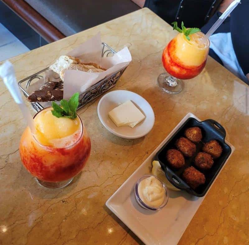 Drinks with fruit garnish, a basket of bread, a plate of butter, and a dish of fried balls with dipping sauce on a marble table.