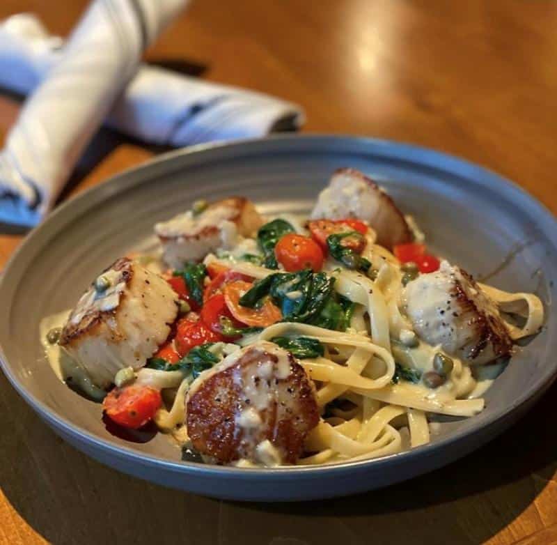 Pasta with seared scallops, cherry tomatoes, and spinach, served on a gray plate. Two rolled napkins in the background.