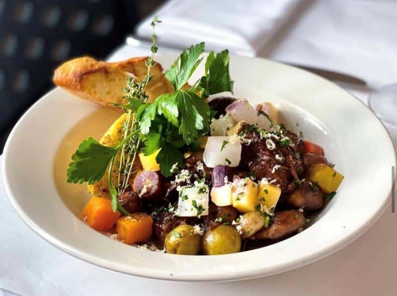 A plate of beef stew with colorful vegetables and herbs, garnished with fresh greens and served with a slice of toasted bread.