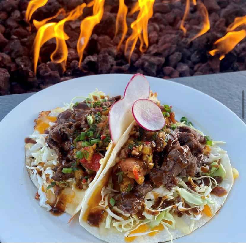Two beef tacos with shredded cabbage, salsa, and radish slices on a white plate. A fire pit with visible flames is in the background.