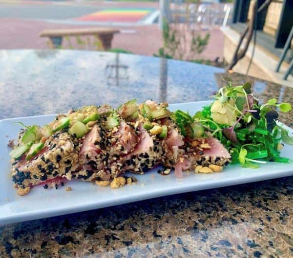 Sliced sesame-crusted tuna with herbs and greens on a rectangular white plate, placed on an outdoor table.