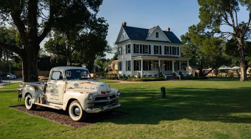 A vintage truck is parked on a grassy lawn in front of a large, two-story house with a covered porch and blue shutters, surrounded by trees.