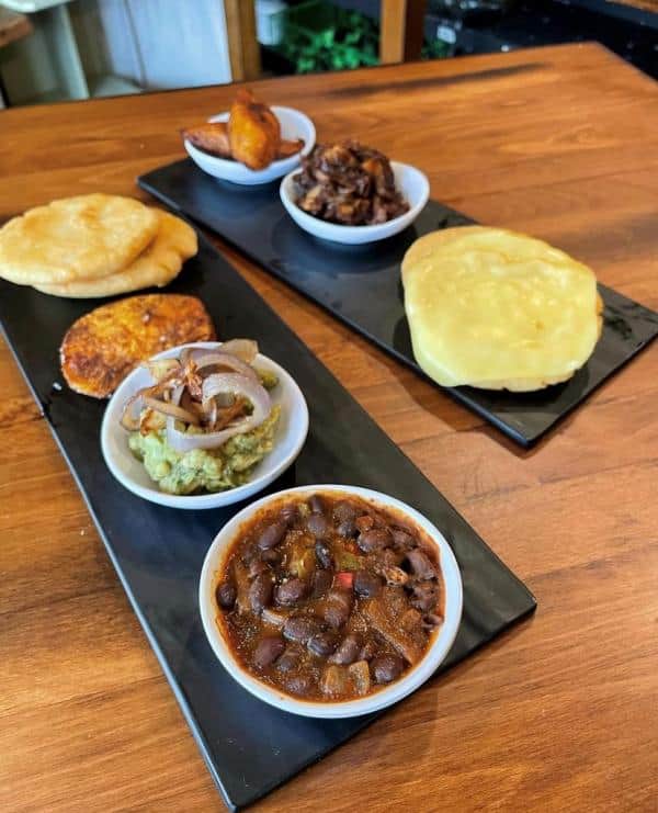 Two rectangular plates with various small dishes: cooked beans, guacamole with onions, fried arepas, plantains, shredded meat, and a large piece of cheese on a wooden surface.