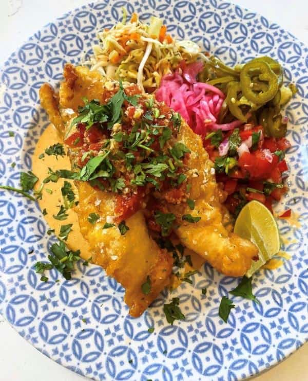 Fried fish on a patterned plate with sliced jalapeños, shredded vegetables, diced tomatoes, pink onions, and a lime wedge.