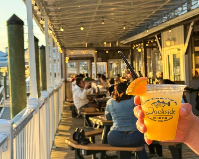 A hand holds a drink in a plastic cup with an orange slice garnish on a sunlit dockside restaurant deck. Diners are seated at picnic tables in the background.