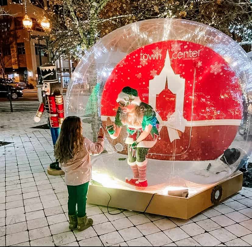 A child high-fives a person dressed as an elf inside a transparent, inflatable snow globe. The setup is outdoors, with a nutcracker statue nearby.