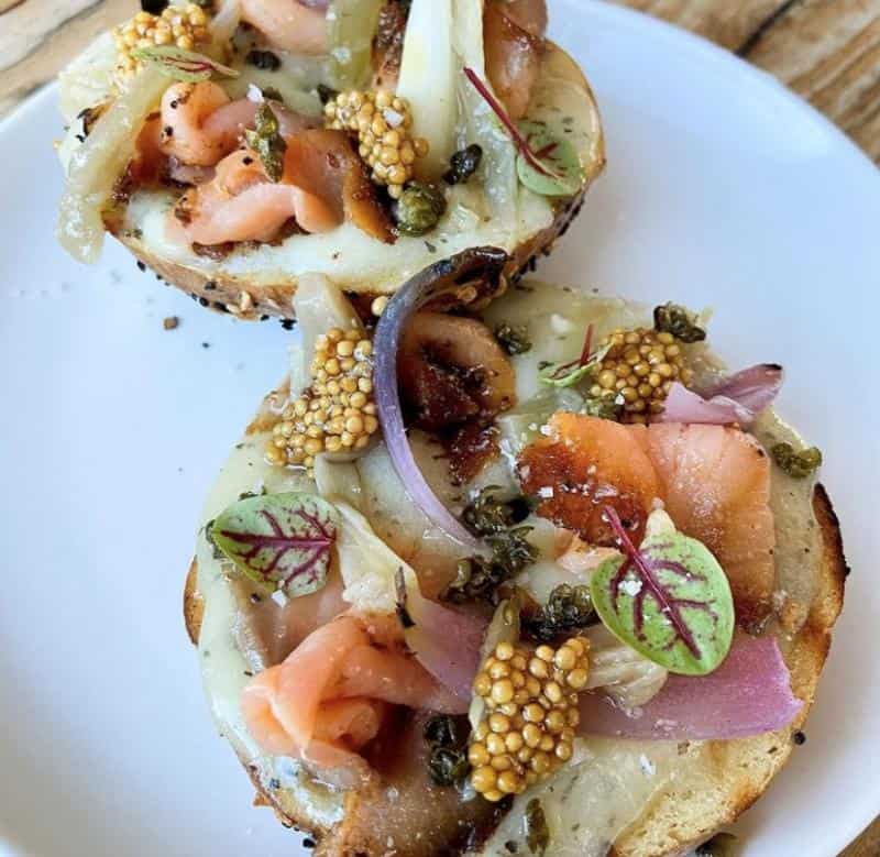 Two bagel halves topped with smoked salmon, pickled mustard seeds, capers, red onion, and microgreens on a white plate.
