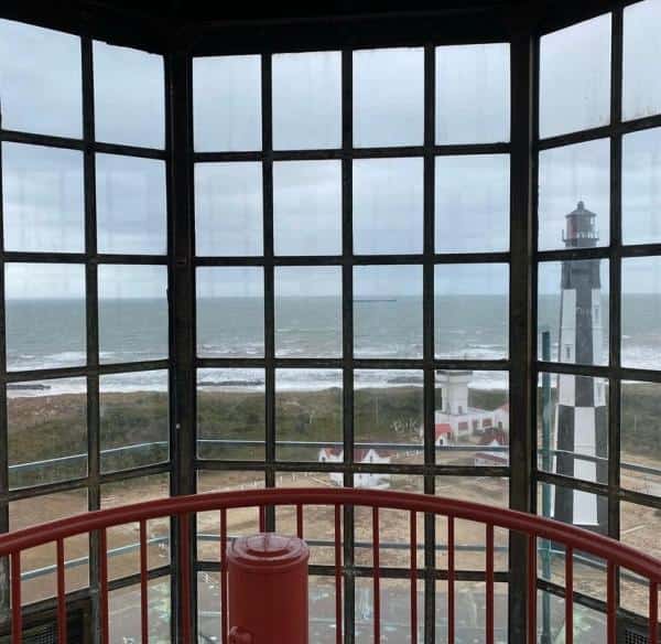 View from inside a lighthouse with glass windows showing an ocean, a distant lighthouse, and coastal buildings.