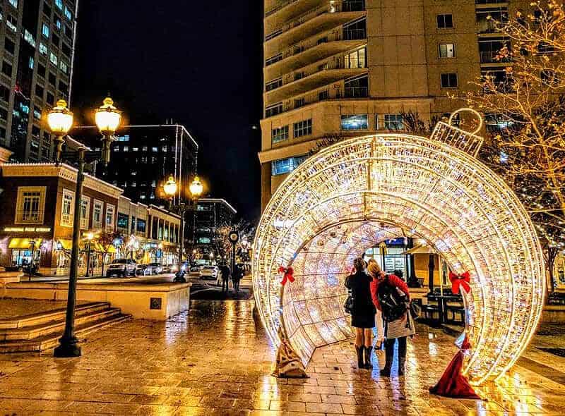 Two people stand under a lit decorative arch in a city plaza at night, surrounded by tall buildings and illuminated streetlights.