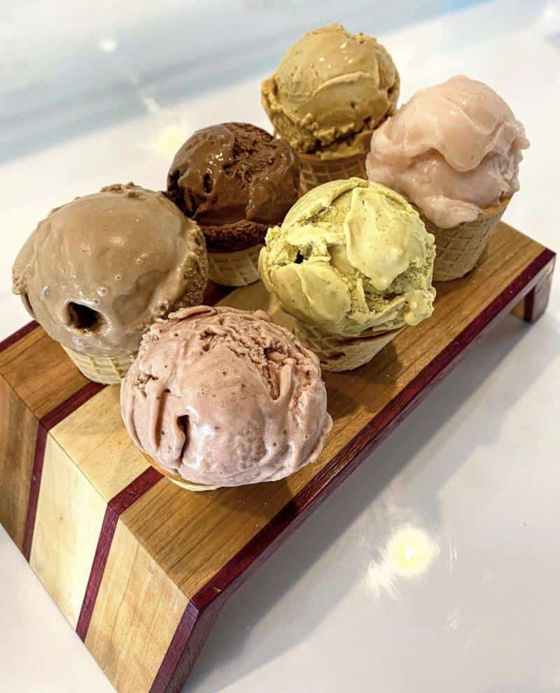 Six ice cream cones with different flavored scoops displayed on a wooden stand.