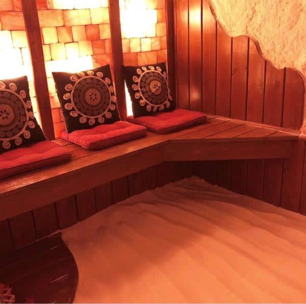 Wooden bench with three decorative pillows and red cushions. The floor is covered in white sand, and the wall has a warm-toned salt block pattern.