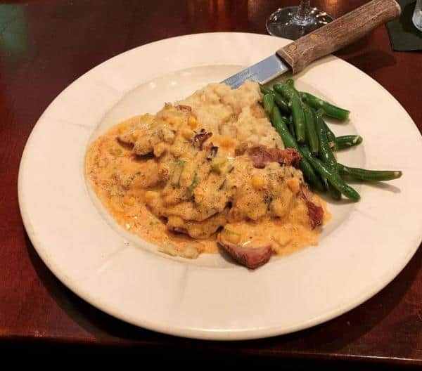 Plate of steak topped with a creamy sauce, served with green beans and mashed potatoes, placed on a wooden table with a steak knife.