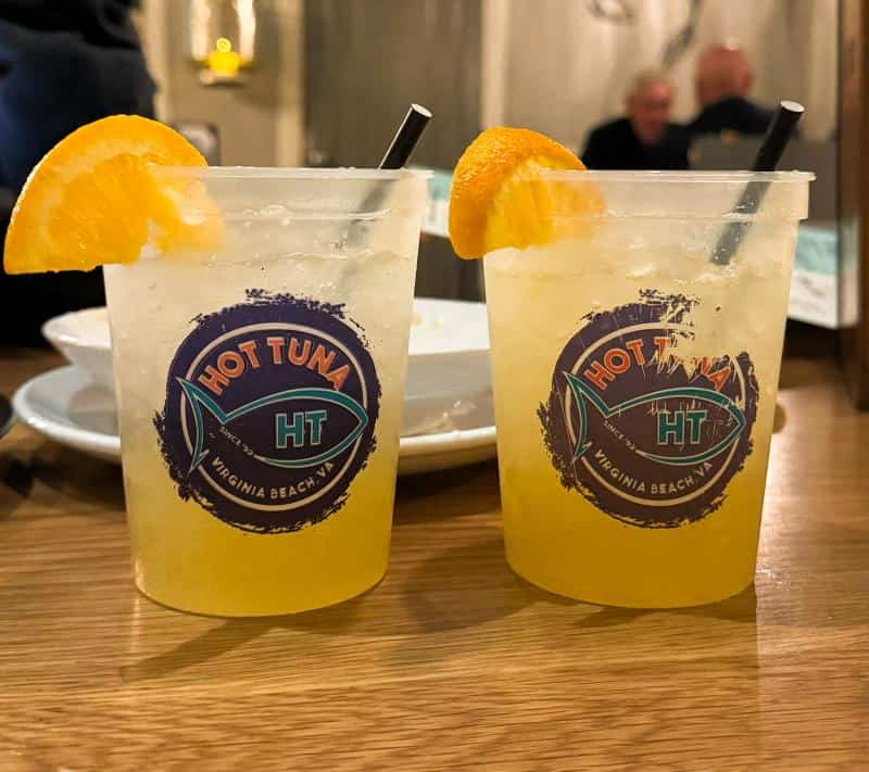 Two plastic cups with a logo reading "Hot Tuna, Virginia Beach" contain a yellow beverage with orange slices and black straws, placed on a wooden table. Plates are visible in the background.