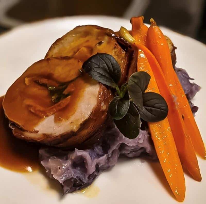 Roasted meat slices with gravy, served on mashed purple potatoes, garnished with glazed carrots and fresh greens on a white plate.