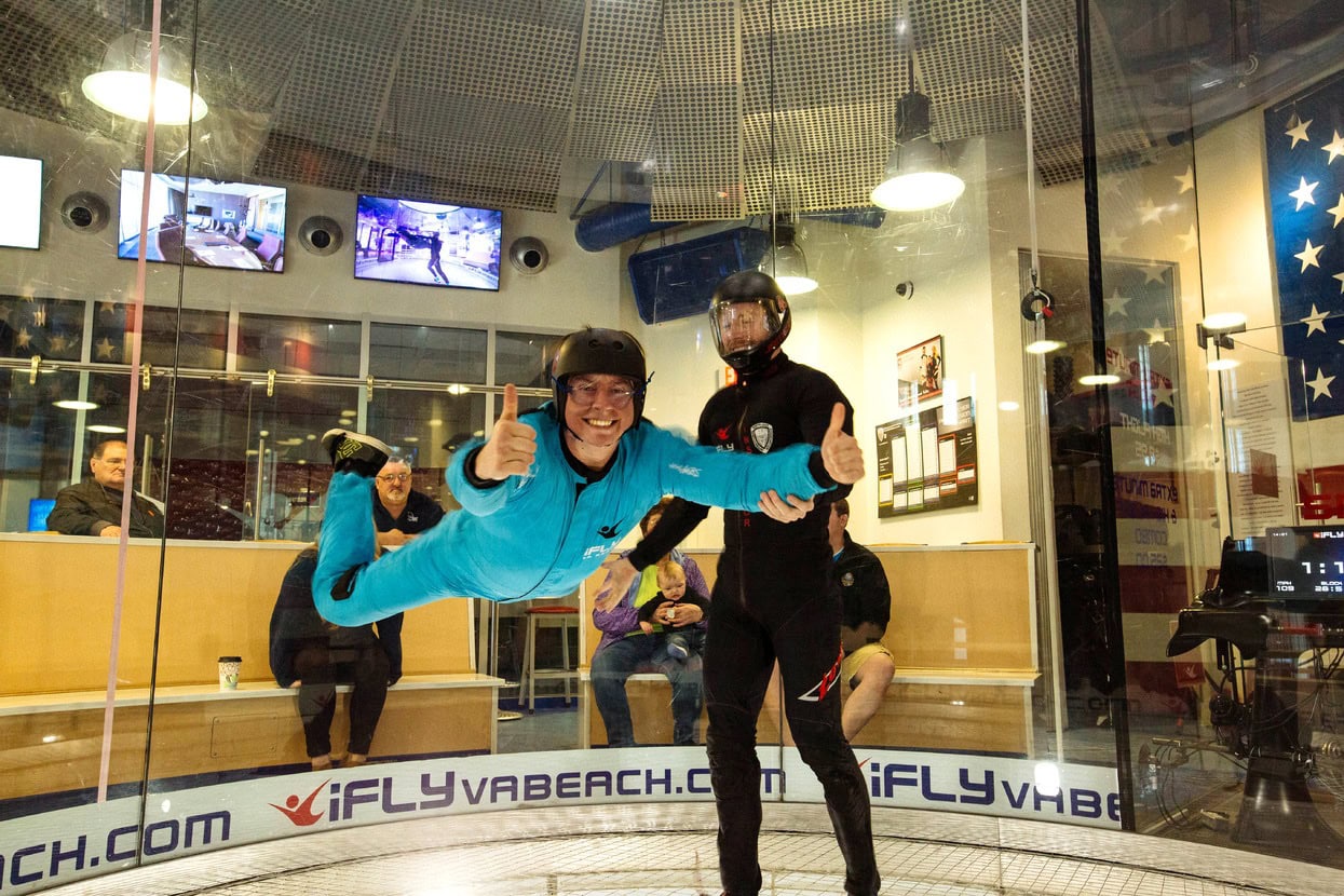 A person in a blue suit is indoor skydiving, supported by an instructor, inside a vertical wind tunnel at iFLY Virginia Beach. Spectators watch from outside the glass.