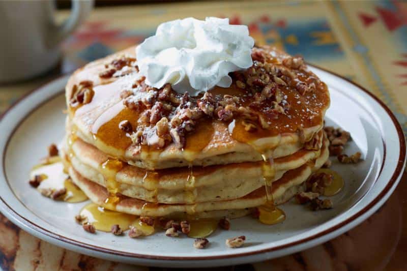A stack of pancakes topped with whipped cream, chopped nuts, and syrup on a white plate.
