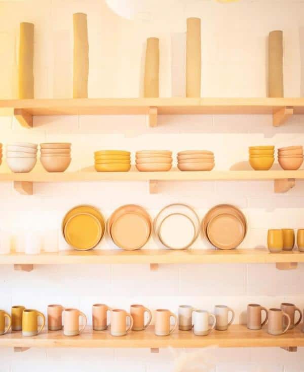 Shelves with neatly arranged pottery, including plates, bowls, and mugs, in various shades of beige and yellow against a light wall.