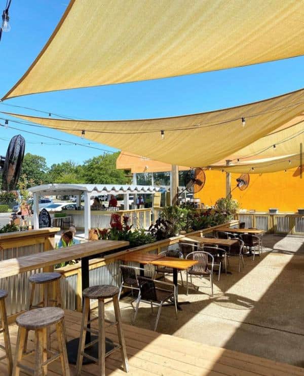 Outdoor patio with wooden bar stools, tables, and sunshade sails at Java Surf. String lights hang above, and plants decorate the area. Bright sunny day with a view of trees and a building in the background.