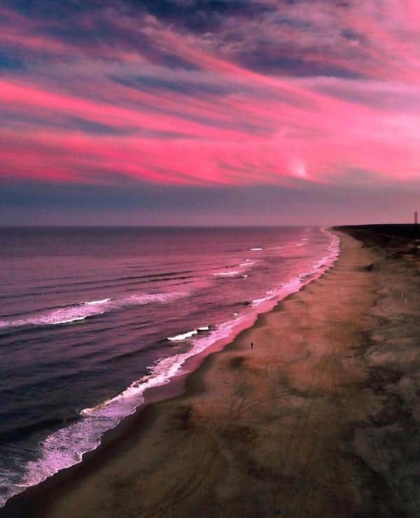 Aerial view of a beach at sunset with pink and purple clouds. Waves gently reach the shore, and a lone figure walks along the sand.
