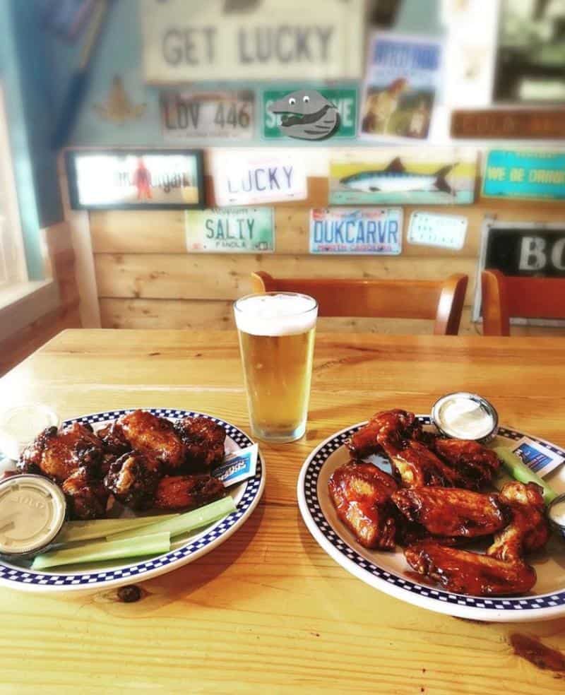 Two plates of chicken wings with dipping sauces and celery on a table, accompanied by a pint of beer. License plates decorate the wooden wall in the background.