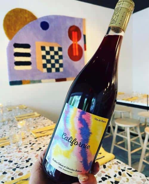 A hand holds a bottle of red wine labeled "Californie" in a modern dining setting with a colorful abstract painting on the wall.