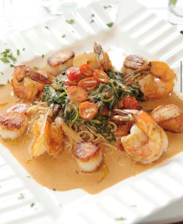 A dish of scallops, shrimp, spaghetti, spinach, and cherry tomatoes in a creamy sauce, served on a white plate.