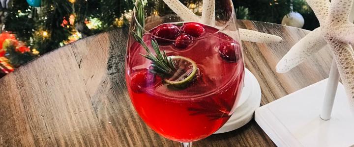 A glass of red cocktail garnished with cranberries, lime, and rosemary on a wooden table near Christmas decorations.