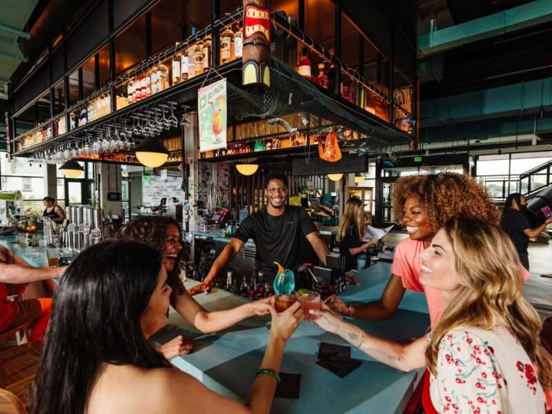 The bar scene at Moxy, featuring four friends toasting with colorful cocktails, while a smiling bartender engages with them in a vibrant atmosphere.