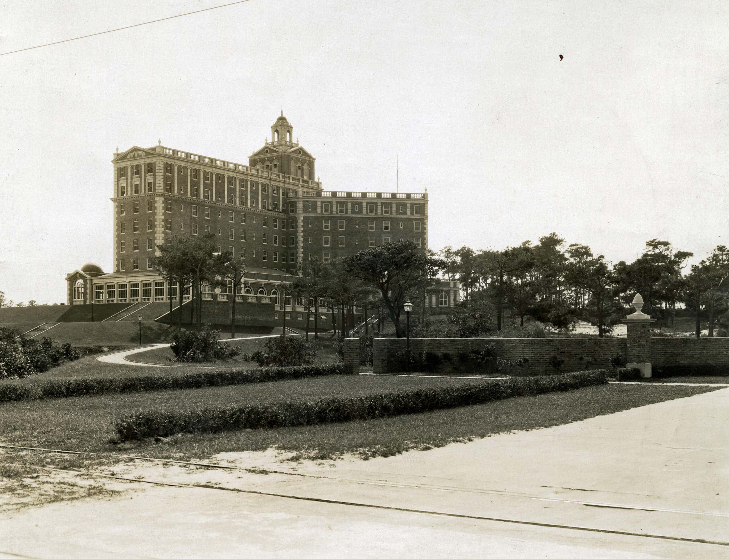 Historic multi-story brick building with a central tower, surrounded by trees and a curved driveway, viewed from a paved road in the foreground.