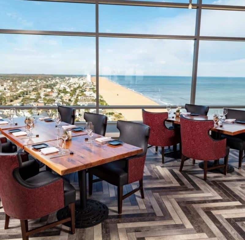 Restaurant with wooden tables and red chairs overlooks a beach and ocean from a high vantage point. Large windows provide a panoramic view.