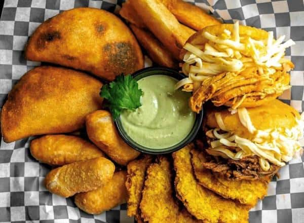 A variety of fried foods including empanadas, plantain slices, and arepas with shredded cheese, arranged around a green dipping sauce on a checkered tray.