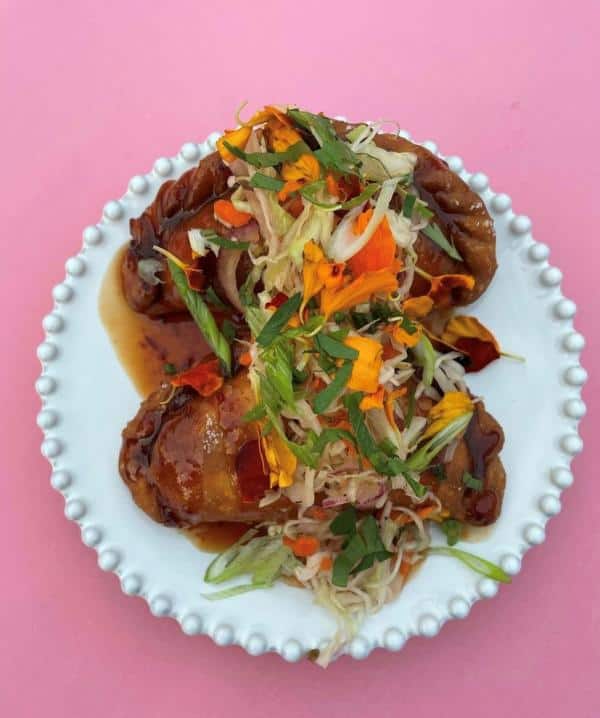 A plate of stuffed fried rolls topped with shredded vegetables and edible flowers on a pink background.