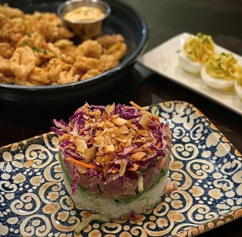 A beautifully plated dish with layers of rice, raw fish, cabbage, and crispy toppings. In the background, a plate of fried calamari and a platter of deviled eggs are slightly blurred.