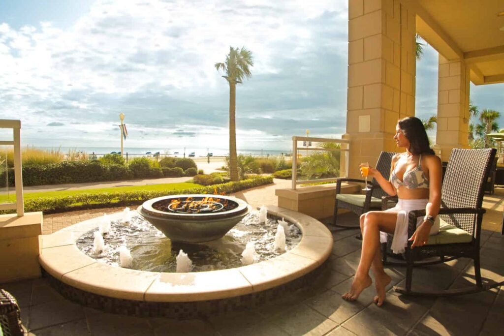 Woman in a swimsuit sits on a patio chair, holding a drink, near a fountain. The background features palm trees and an ocean view under a cloudy sky.