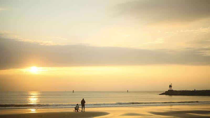 Two people stand together on a sandy beach, looking at the ocean during sunset, with a lighthouse in the distance.