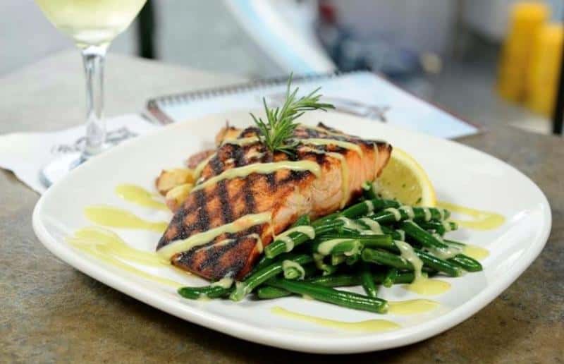 Grilled salmon on green beans topped with sauce and garnished with rosemary, served with sliced lemon. White plate on a table with a glass of white wine in the background.