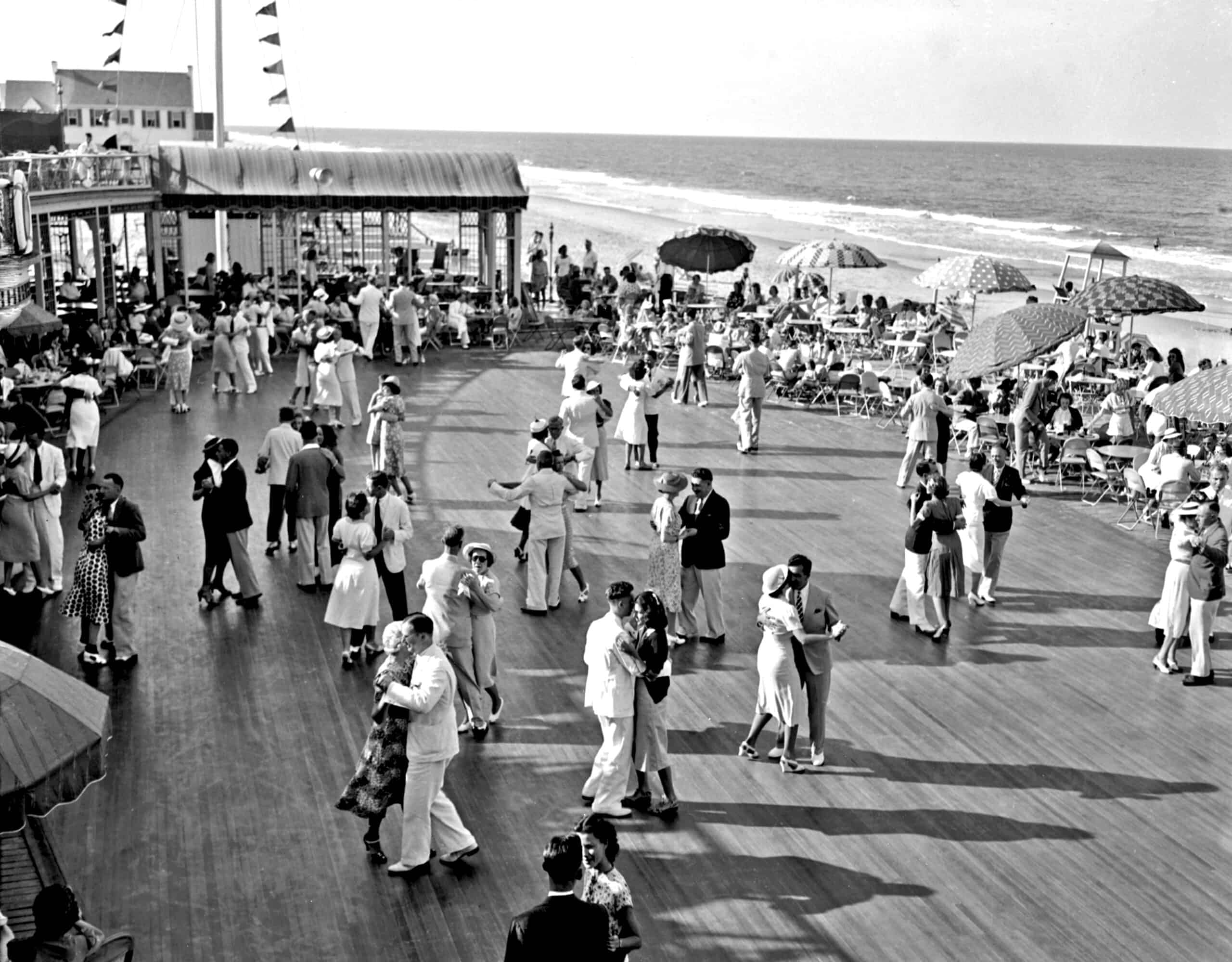 People dance and socialize on a bustling boardwalk by the beach, with parasols and seated onlookers in the background.