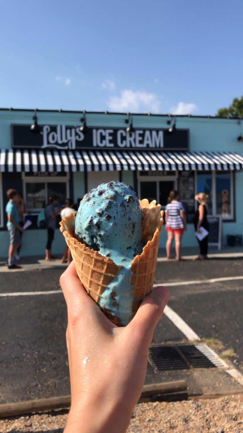 A hand holds a waffle cone with blue ice cream outside Polly's Ice Cream shop. People are lined up under the awning.