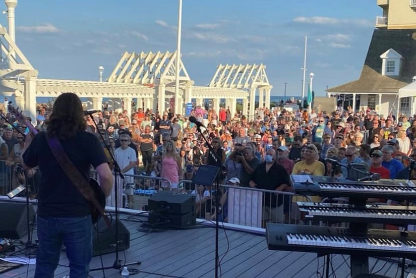 A musician performs on stage in front of a large crowd at an outdoor concert, with a lively atmosphere and blue sky in the background.