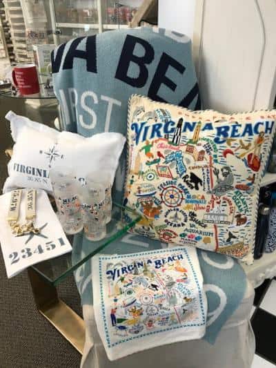 Assorted Virginia Beach-themed souvenirs including pillows, a towel, glasses, a lanyard, and a coaster displayed on a shelf.