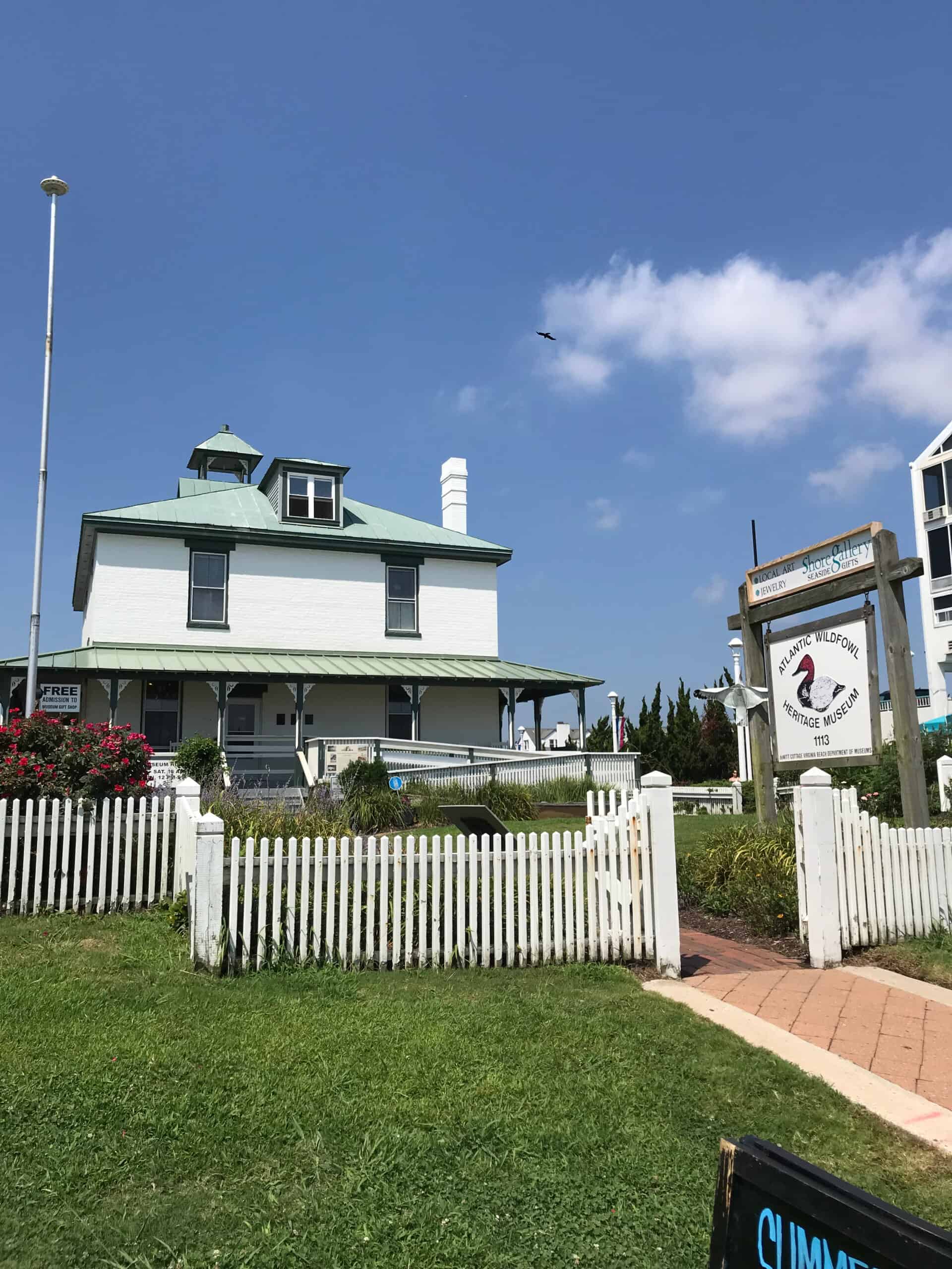 Historic white lighthouse museum with a green roof, surrounded by a white picket fence, under a clear blue sky. A signboard is visible at the entrance.